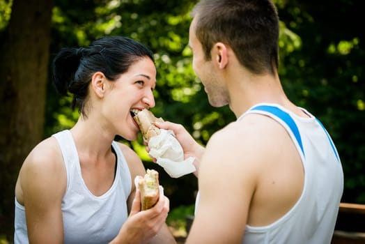 Young couple eating after sport training outdoor in nature - man feeds woman