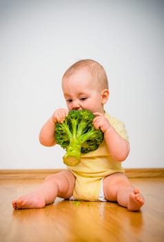 Cute baby eating broccoli sitting on floor indoors