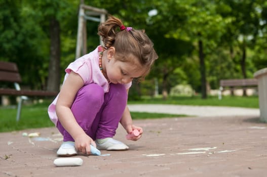 Small cute girl drawing with chalk on sidewalk, trees in background