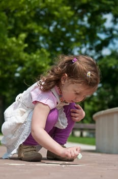 Small cute girl drawing with chalk on sidewalk, trees in background