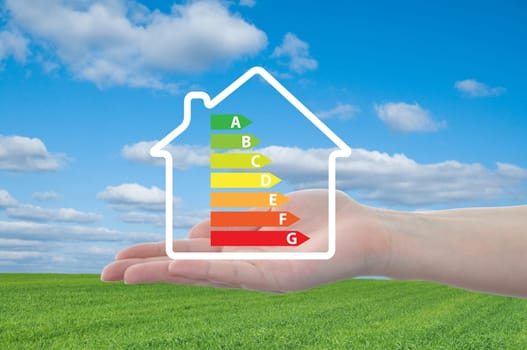 house and energy efficiency graph on hand. blue sky and meadow in background.