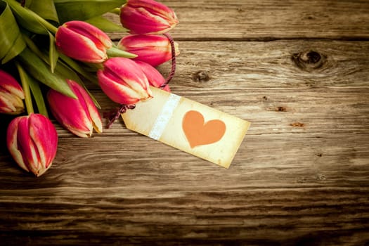 Vintage Valentines or anniversary gift with an aged yellow gift tag or label with a red heart attached to a bouquet of fresh red tulips on a weathered rustic wooden background with copyspace