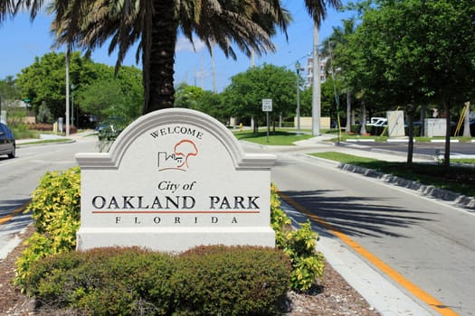 OAKLAND PARK, FLORIDA - MAY 7, 2013: Cement sign with buildings and tree graphic welcoming people to the city of Oakland Park, Florida on a tropical foliage street median on a sunny blue sky day.
