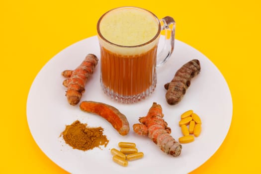 Turmeric juice beverage cup, three fresh and raw conventional rhizome roots, one turmeric root partially peeled, standardized extract capsules and tablets, along with some dry ground turmeric powder.