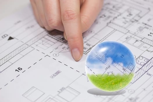 Finger from an unknown person is showing at the plan of the house. Blue sky, blueprint and green grass are in the glass ball.