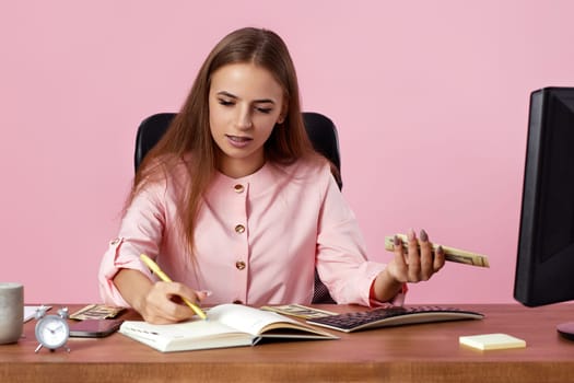 accountant woman holding money and planning financial statistics while sitting on chair at desk on pink background