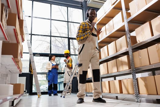 African american man in headphones dancing in parcels storage room. Happy storehouse young worker in good mood listening to music while working in post office service warehouse