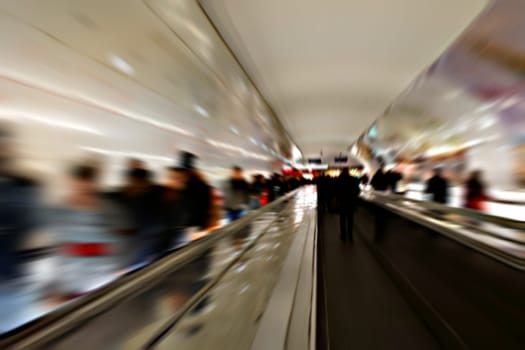 The railing of an escalator in a subway tunnel with passing people photographed with long exposure.