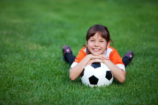Outdoor photo of cute little girl leaning on soccer ball in green grass