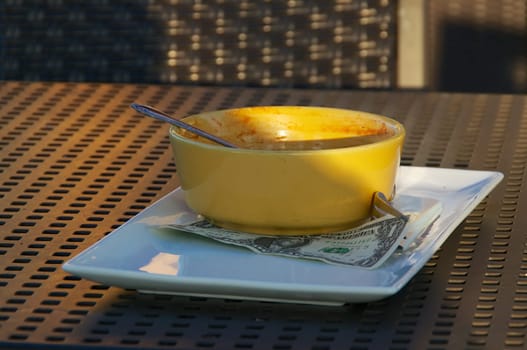 An empty yellow soup bowl sits on an outdoor cafe table with money tip on tray.