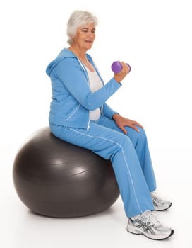Full length studio photo of mature woman seated on balance-ball lifting dumbbell on white background.