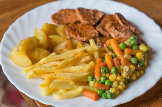 Steak with fries and fried potatoes,with corn,carrots and peas. Served on a white plate.