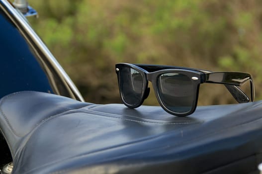 A pair of generic wayfarer sunglasses are on top of leather motorcycle seat. NOTE