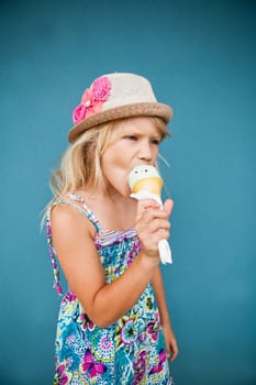 Cute young girl eating vanilla ice cream in cone outside against blue wall background