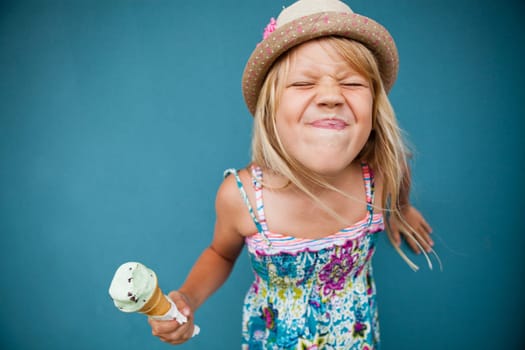 Cute young girl with funny expression holding ice cream cone outside against blue wall background