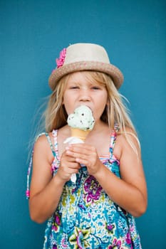 Cute young girl eating vanilla ice cream in cone outside against blue wall background