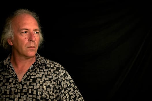 older white male with grey hair and blue eyes wearing patterned shirt, looking left into darkness.