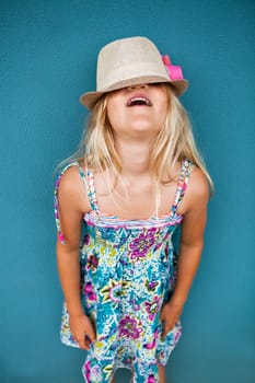 Portrait of stylish cute young girl against blue wall background with hat covering eyes and funny expression