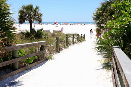 SIESTA KEY, FLORIDA - MAY 9, 2013: People entering very wide and colorful Siesta Beach on Siesta Key through a sand covered wood walkway with wood railings surrounded by foliage on a sunny day.