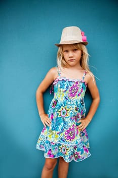 Portrait of serious looking cute young girl wearing stylish hat outside against blue wall background