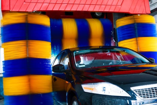 black car being washed in automatic car wash,visible blue and yellow rotating brushes.