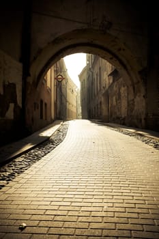 a fragment of the old town cobbled street in the sunlight in the down,empty street visible arch of the gate above the street.