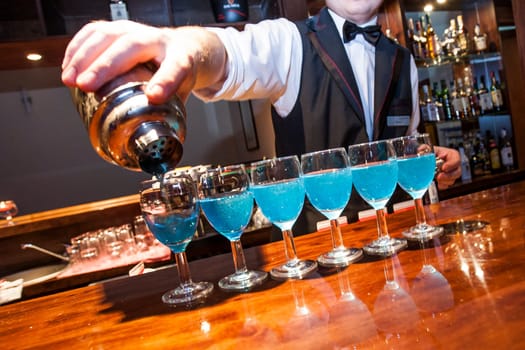 Barman, bartrender, pouring blue coloured drinks from the shaker to the row of glasses  on the bar counter, visible fragment of the hand, shaker and  barman. No need model relase.