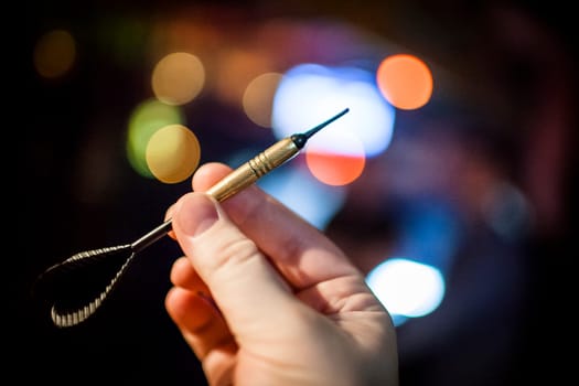 In the photo visible dart with a black feather, visible hand, fingers holding a dart, spots of light in the background.