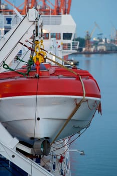 White and red lifeboat, visible ropes and elements of the lift, in the background port, cranes and sea.
