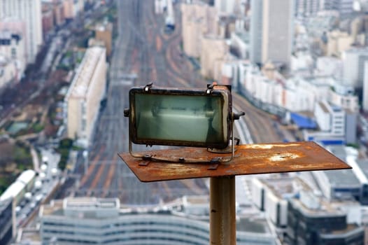 The photograph of a rusty spotlight on a high-rise building with rails in the background.