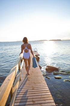 Attractive woman in white bathing suit walking on wooden pier in nordic country during summer