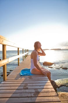Attractive woman in white bathing suit sitting on wooden pier in nordic country during summer