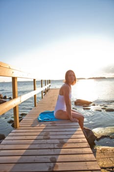 Attractive woman in white bathing suit sitting on wooden pier in nordic country during summer