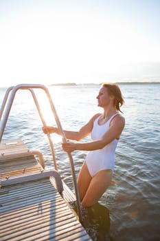 Attractive woman in white bathing suit climbing up ladder on to wooden pier in nordic country during summer