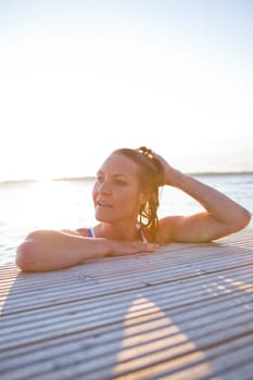Attractive woman in white bathing suit resting at edge of wooden pier in nordic country during summer