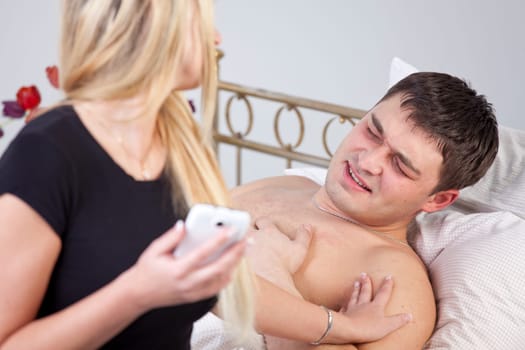 Sick man with ache on bed, worried woman holding phone at his side.