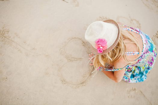 Young girl with summer dress and stylish hat drawing heart shapes in sand on beach with finger