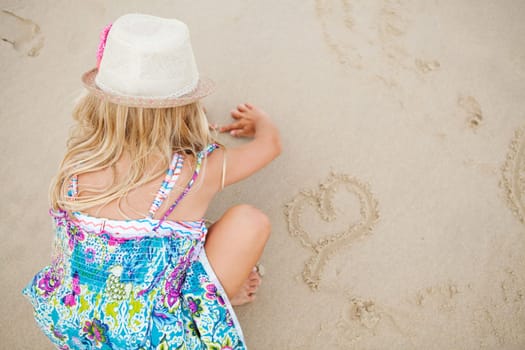Young girl with summer dress and stylish hat drawing heart shapes in sand on beach with finger