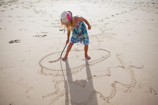 Young girl with summer dress and stylish hat drawing in sand with stick on beach