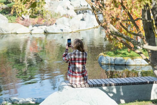 Woman in red plaid shirt enjoying nature standing in Japanese Garden taking photo on smartphone