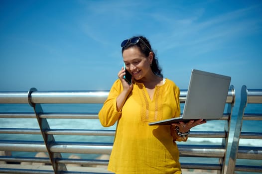 Beautiful smiling woman freelance entrepreneur talking on mobile phone, negotiating with business partners, holding laptop and online working outdoor. Freelance occupation. Distance work