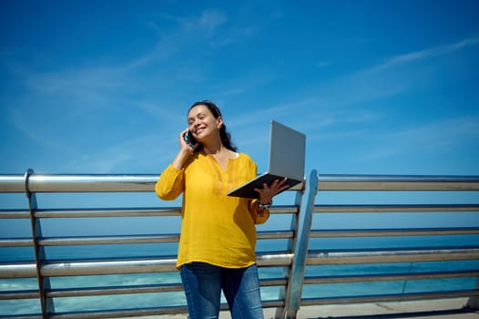 Waist up portrait of a middle aged confident successful freelance worker, manager, a charming business woman holding laptop and smiling broadly while talking on smart mobile phone outdoor. Copy space