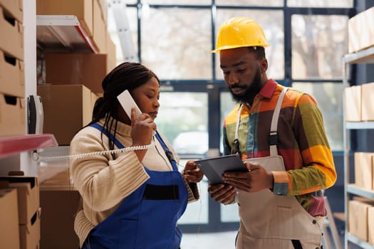 Warehouse manager calling in logistics office and asking about inventory report. African american storehouse workers team looking at stock information on tablet and talking on telephone