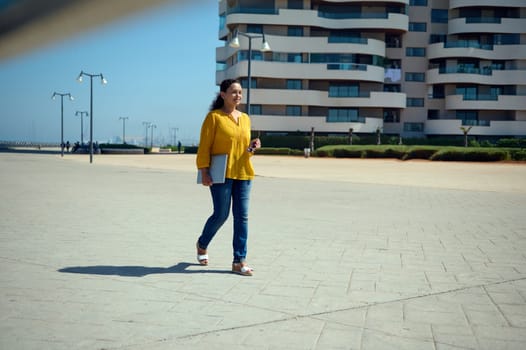 Full length portrait of a young adult woman dressed in blue denim and yellow casual shirt, strolling the urban street with a laptop in hands, smiling broadly while going to the office. People and job