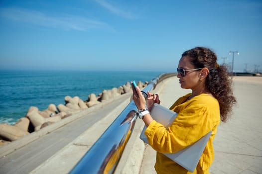 Side portrait of a beautiful curly haired brunette, dressed in yellow casual shirt, holding laptop and checking mobile app on her smart phone, standing by a metal fence in a promenade. Copy ad space