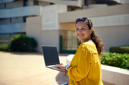 Close-up smiling middle aged multi ethnic confident business woman in casual attire, smiling looking aside, using a laptop with black blank mockup screen. Copy advertising space for mobile application