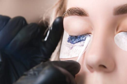 Close-up portrait of a woman having eyelash tinting procedure. A handyman wearing gloves removes excess paint with a cotton swab.