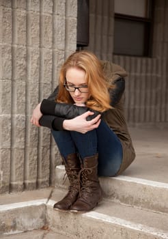 Outdoor photo of young teenage girl seated on concrete steps, hugging knees, distraught facial expression.