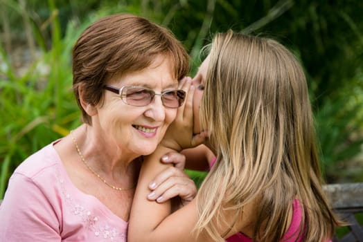 Little girl whispering her grandmother a secret to ear - outside in garden
