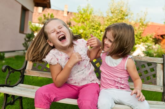 Two small girls (sisters) larking in backyard sitting on bench